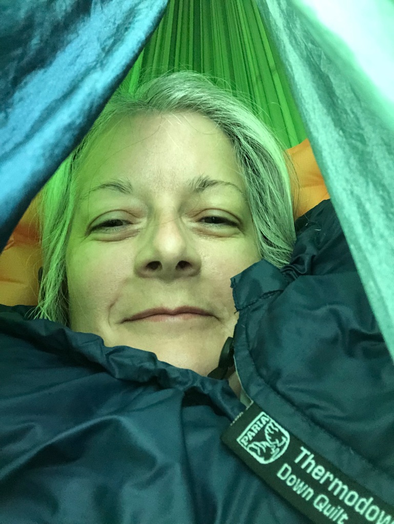 The photo is of Ruth's smiling face surrounded by her green and blue hammock. She is covered by her Paria Thermodown Quilt.