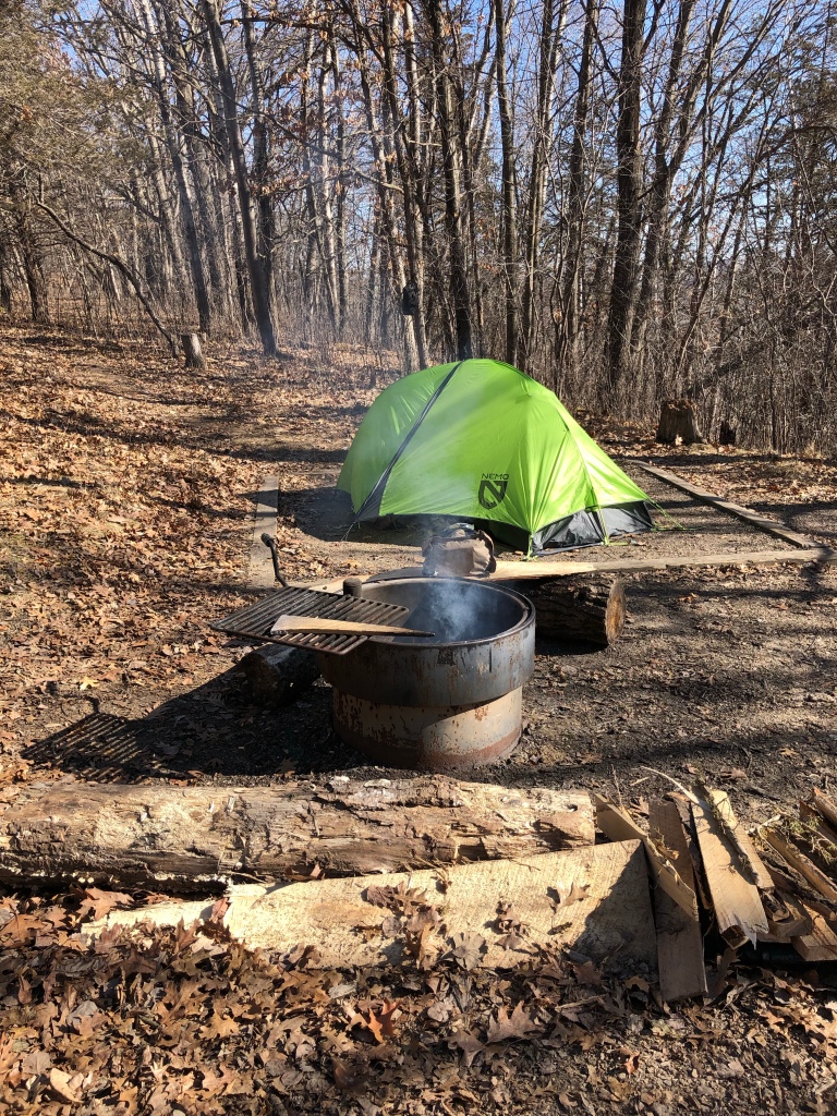 The image shows Ruth's bright green tent sitting in the tent pad which is bordered by a wood frame set into the ground. The campfire ring is in the foreground with a wisp of grey smoke rising from inside. The trees surrounding the image are bare of trees. It's early in the day with a blue sky seen between the trees.