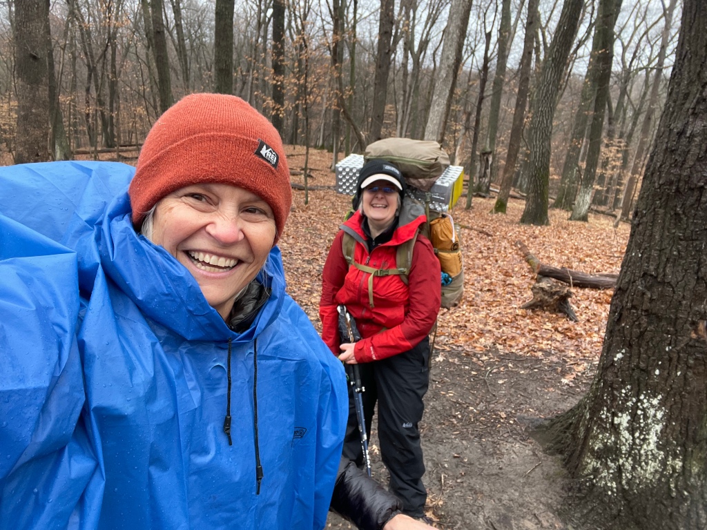 Lynae is in the front of the picture smiling broadly while wearing a bright blue rain poncho and clay colored stocking cap. Ruth is standing behind her, laughing in her red rain jacket with her backpack on. The are standing in a wooded area covered in fallen leaves.